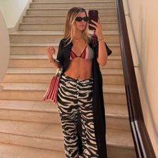 Sofia Richie Grainge stands at the bottom of a staircase taking a selfie in a mirror, wearing zebra print trousers with a multicoloured, striped bikini top and red flip-flops. 