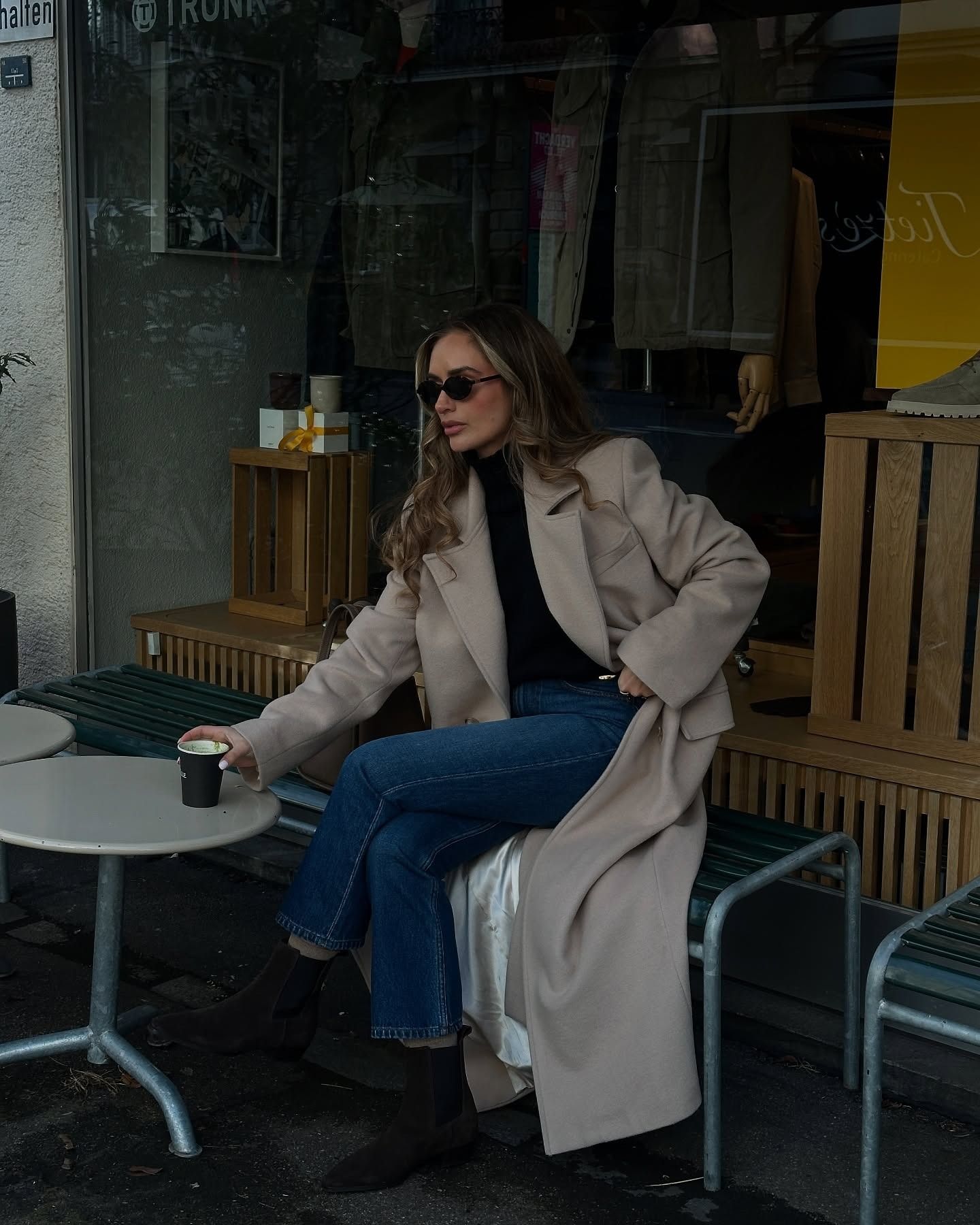 Woman wears black turtleneck jumper, beige coat, blue jeans and black boots