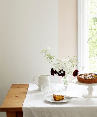 A boho dining room with a light wooden table with a white linen tablecloth, a plate with a slice of yellow cake, a white cakestand with pie on it, a glass vase of purple and white flowers, and a window behind it