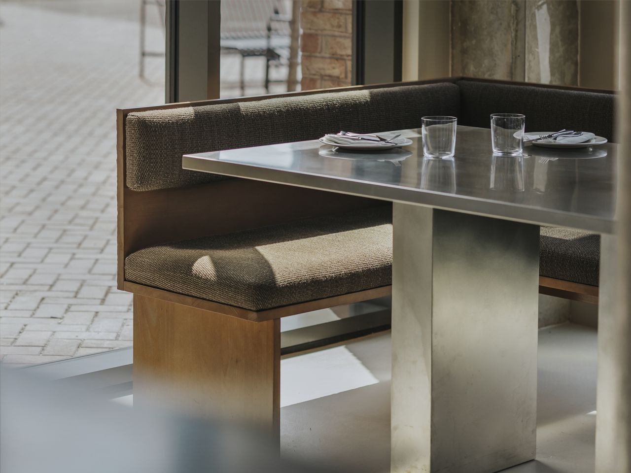 A restaurant with brutalist interiors features wooden upholstered banquettes with warm, brown fabric matching with matte chrome tables with a geometric shape.