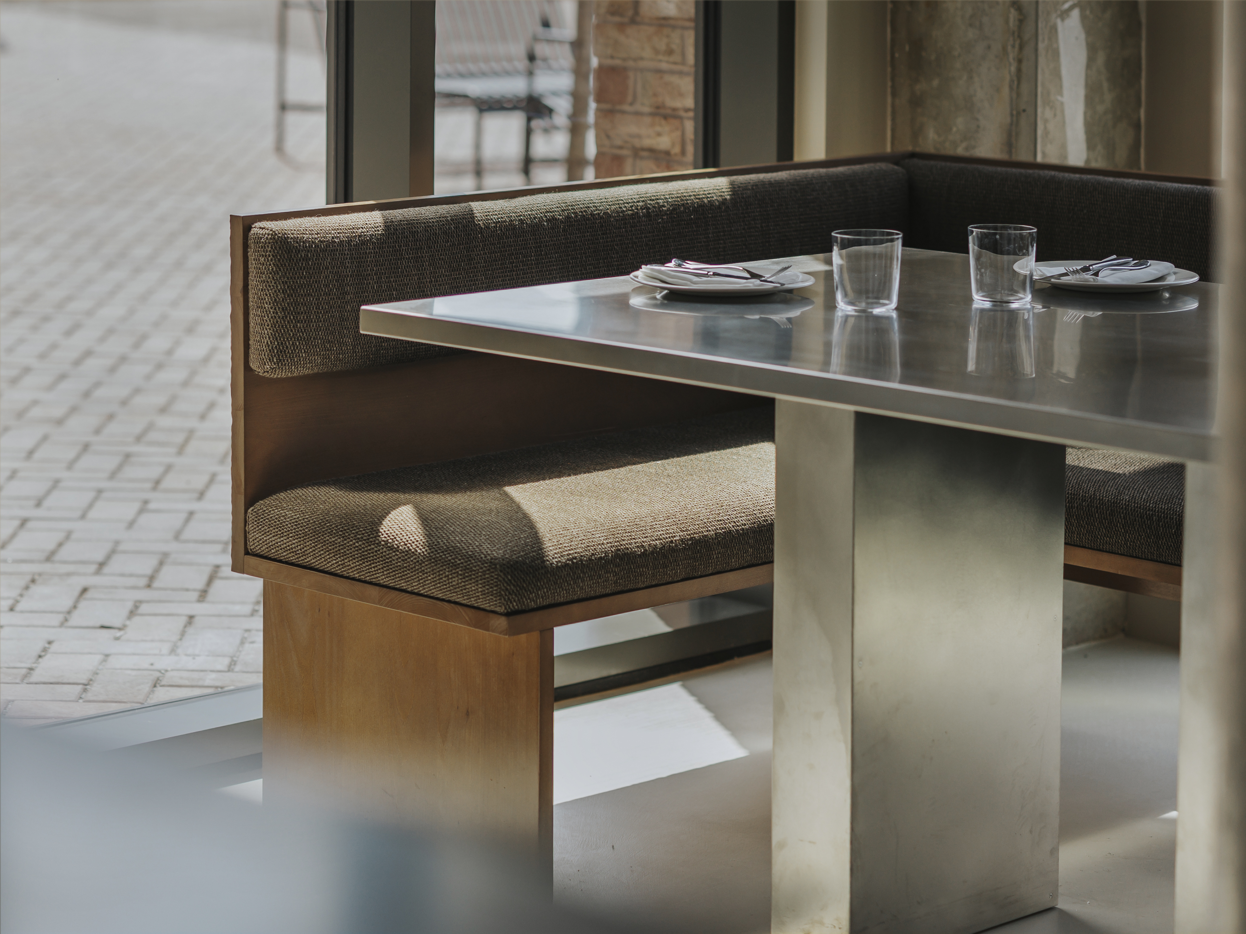 A restaurant with brutalist interiors features wooden upholstered banquettes with warm, brown fabric matching with matte chrome tables with a geometric shape.