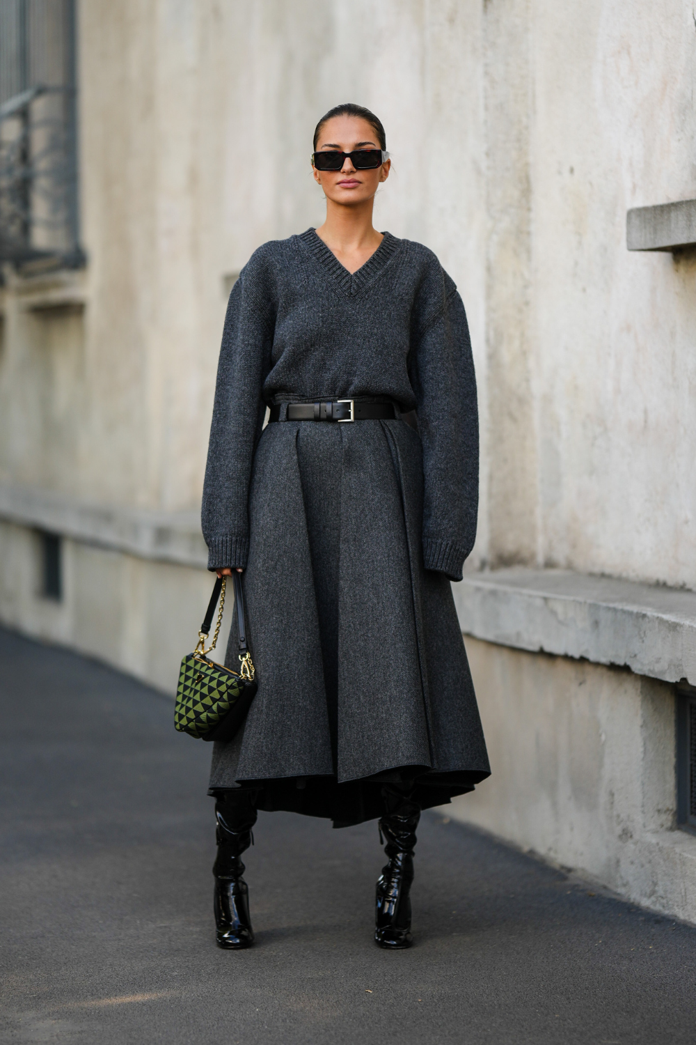 Gabrielle Caunesil wears black sunglasses from Prada, a dark gray wool long sleeves / midi long dress from Prada, a black shiny leather belt from Prada, black shiny varnished heels waders / knees boots, a black and green triangular print pattern Triangle handbag from Prada, outside Prada, during the Milan Fashion Week