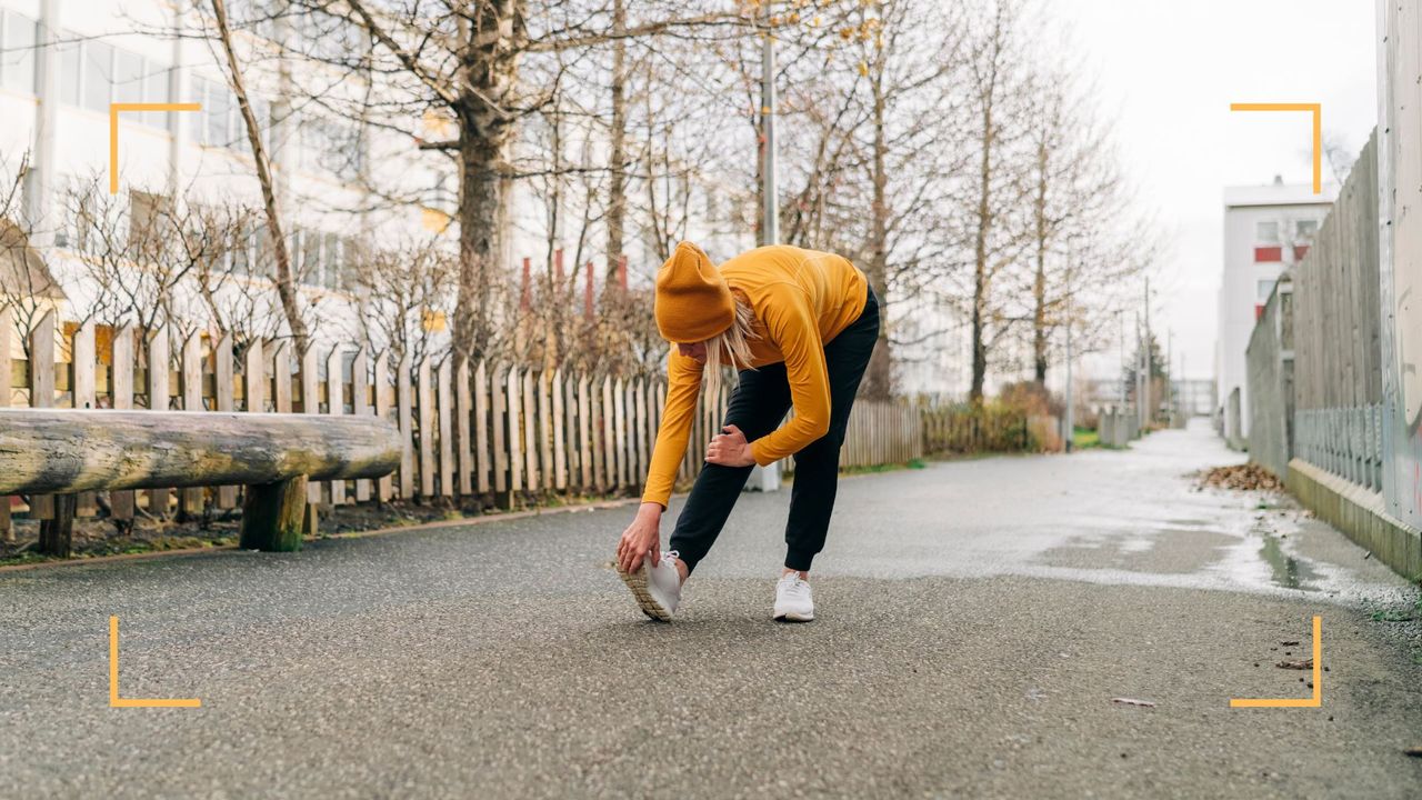 Woman wearing winter running clothes stretching her hamstrings with headphones on outside, before starting one of the best running apps