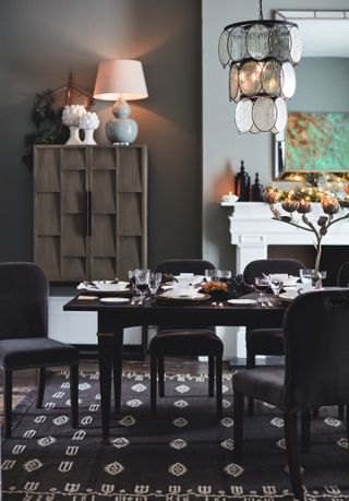 dining room with grey walls, fireplace, tallboy, table lamp, pendant light, black table, grey upholstered chairs, black rug