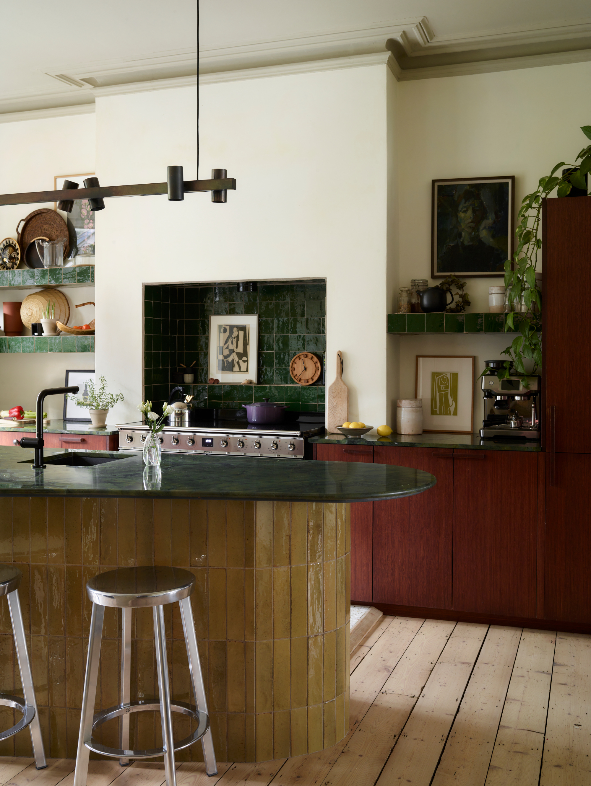 Kitchen with dark red wood cabinets, yellow tiled island, dark green stone countertops and zellige splashback tiles, chrome bar stools, and shelving lined with objects