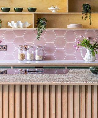 A kitchen with wooden shelves with bowls and plates on, a pink hexagonal splashback, a white counter, and a kitchen island with a brown stone countertop and light wooden panels