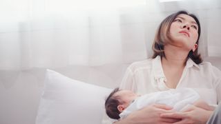 A young Asian woman holds her sleeping newborn baby as she rests her head behind her, looking sad.