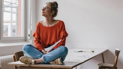 A woman sits crossed-legged on a desk, looking out the window pensively.