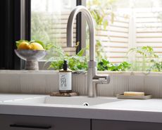 A kitchen sink with a brushed steel faucet, a white quartz countertop, a bar of soap, and a window with a view into a garden