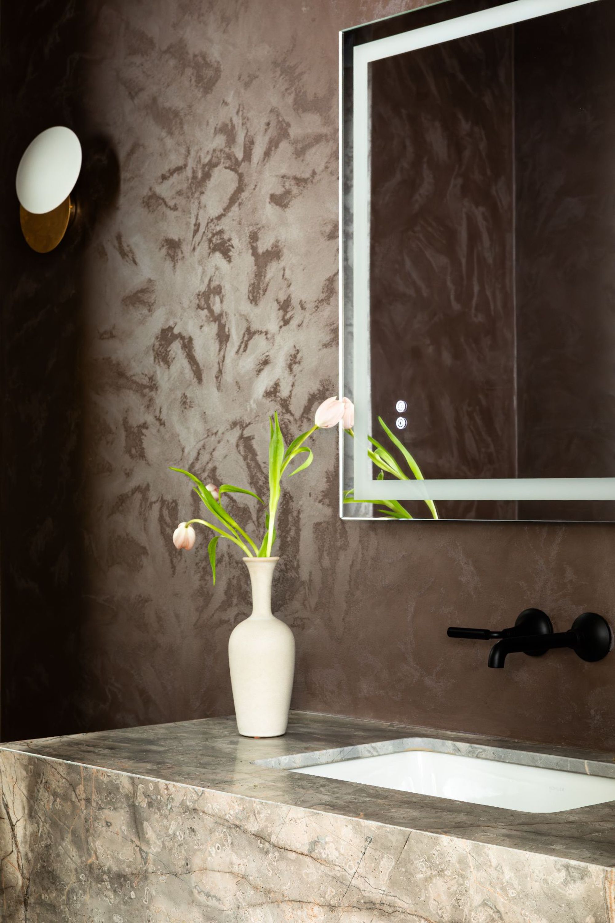 A mid tone marble sink with black hardware and a large mirror above, the walls are plastered in a warm, dark brown colour with a wall lamp in the top left corner.