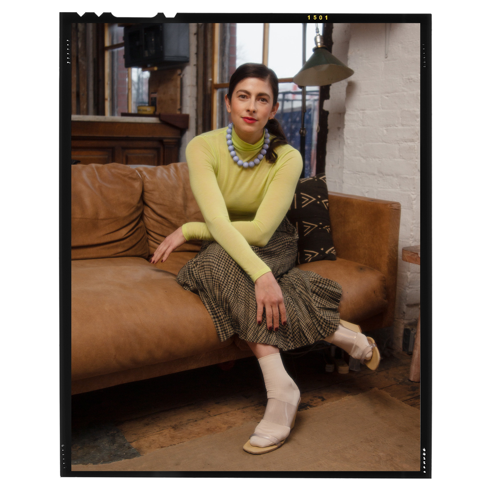 a portrait of erin allweiss sitting on a brown leather couch wearing a green turtleneck, brown plaid skirt, white socks, clear heels, and a blue necklace