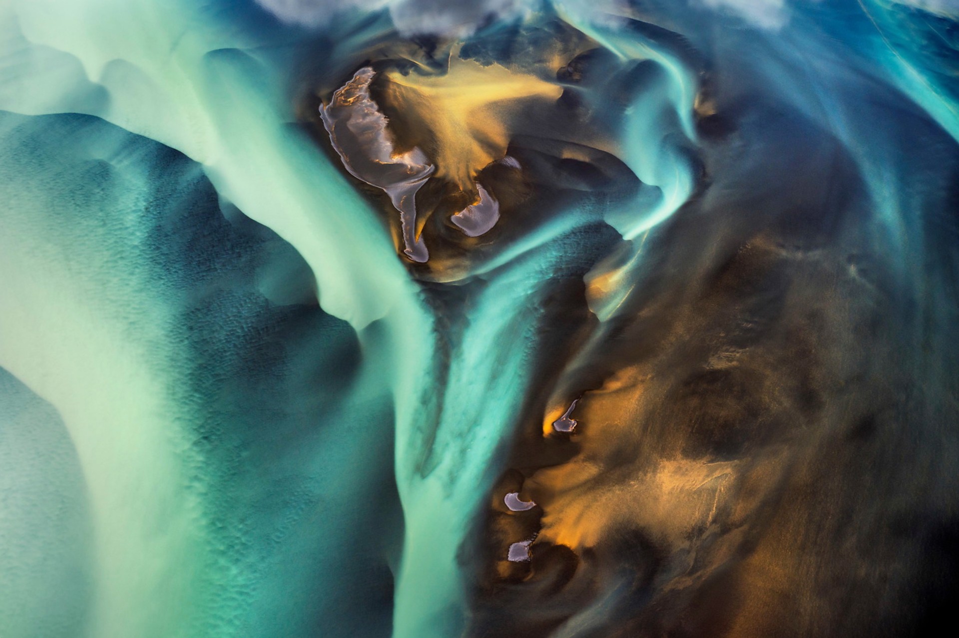 Aerial view of Iceland's Hv&iacute;ta river, its blue and gold meltwater channels folding across dark volcanic sediment like marbled paint.