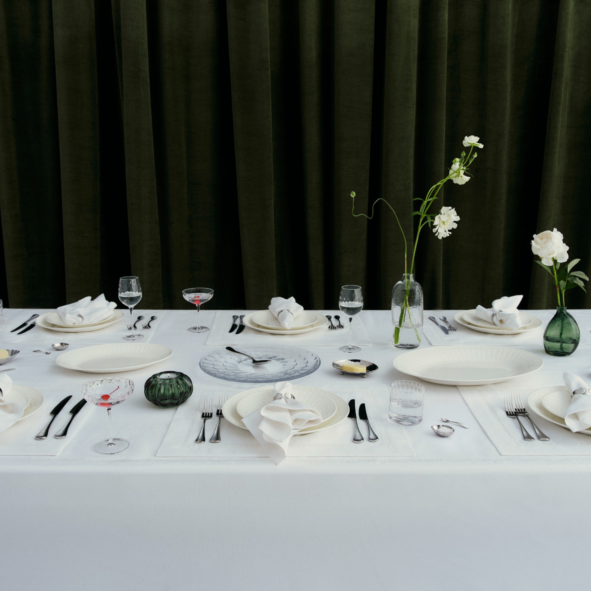A dining table set with ictoria Beckham's tableware from her collection with Westwing set against a dark green velvet curtain
