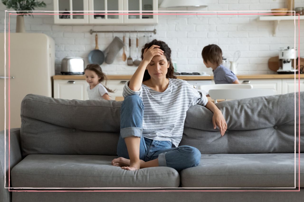 A tired woman sat on a sofa while two children run around in the background