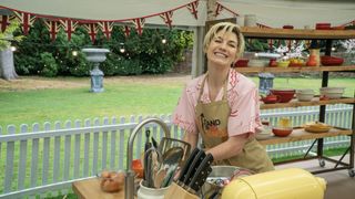 Jodie Whittaker in a pink shirt and brown Stand Up To Cancer apron stands at her work bench in The Great Celebrity Bake Off for Stand Up to Cancer