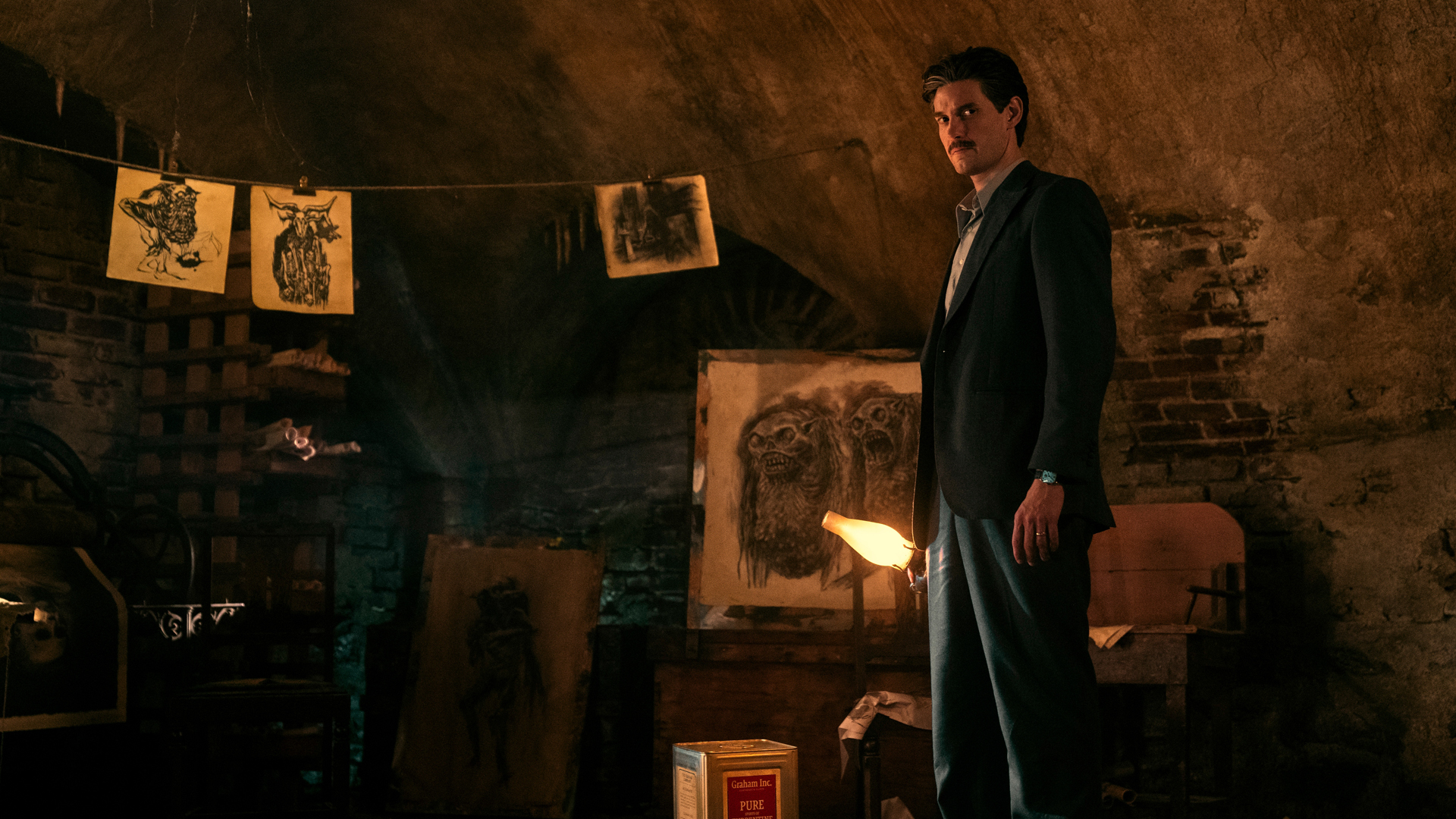 Will Thurber stands in a fire-lit room with a lantern in hand next to artwork in Pickman's Model on Netflix