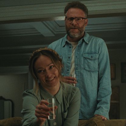 olivia wilde as angela and seth rogen as joe standing behind her as she sits on a couch and cheers in a still from the invite