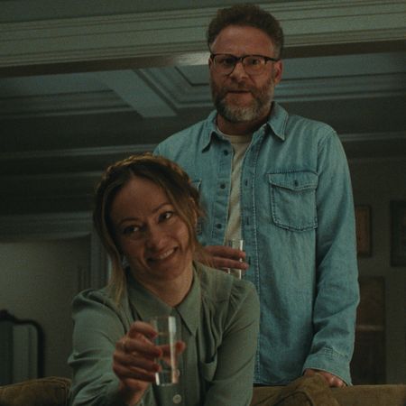 olivia wilde as angela and seth rogen as joe standing behind her as she sits on a couch and cheers in a still from the invite