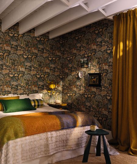 a bedroom with exposed ceiling beams, wallpapered walls, a large bed with colorful pillows and a blanket, and long drapes against the wall