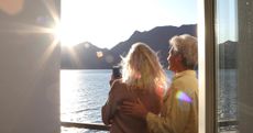 An older couple look out from railing above lake, with mountains in the distance, at sunrise.