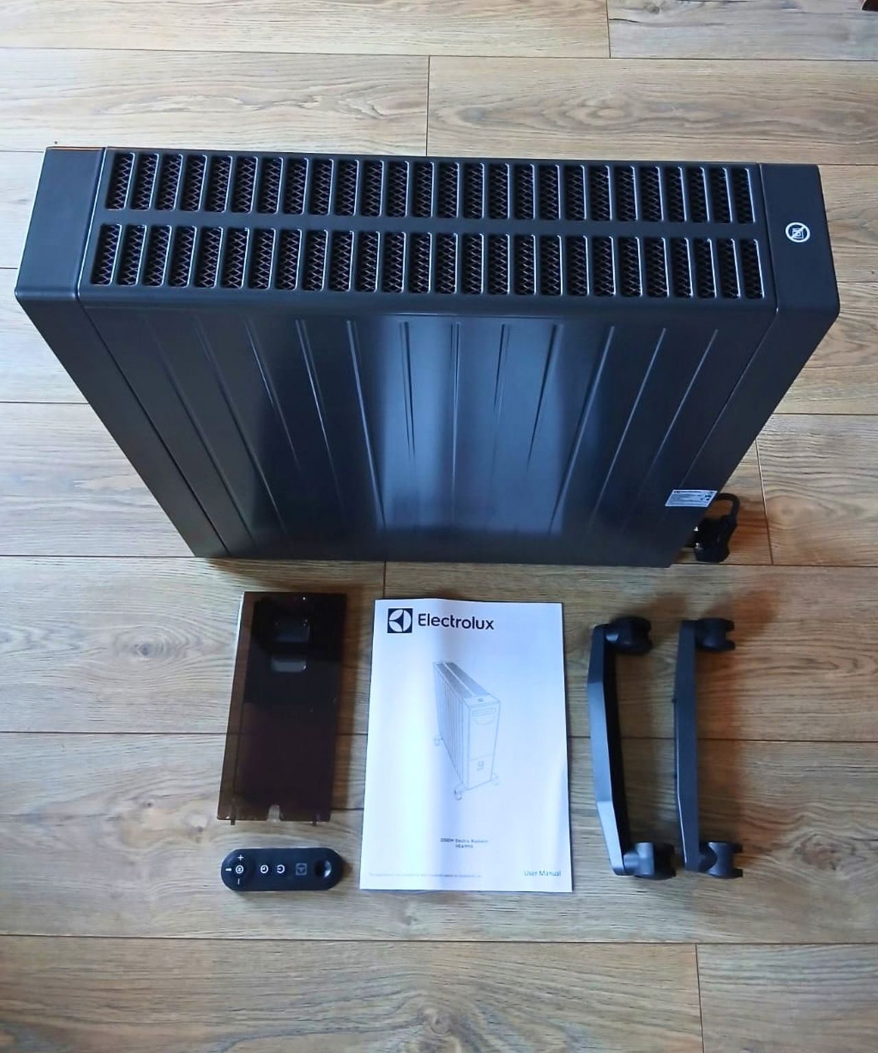 Unboxed black heater laid flat with its cover panel, remote, user manual, and two castor legs.