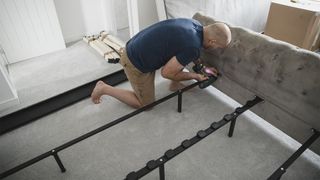 A man kneels on the floor as he attaches the metal poles of his metal bed frame to the headboard