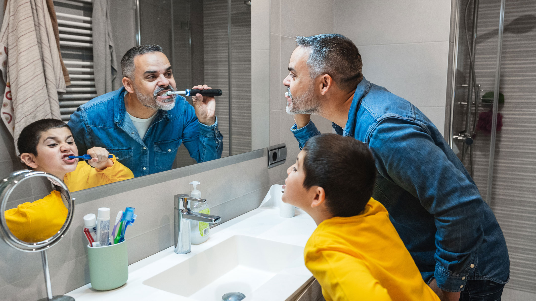 A picture of a father and young son brushing their teeth with electric toothbrushes while standing in front of a bathroom mirror