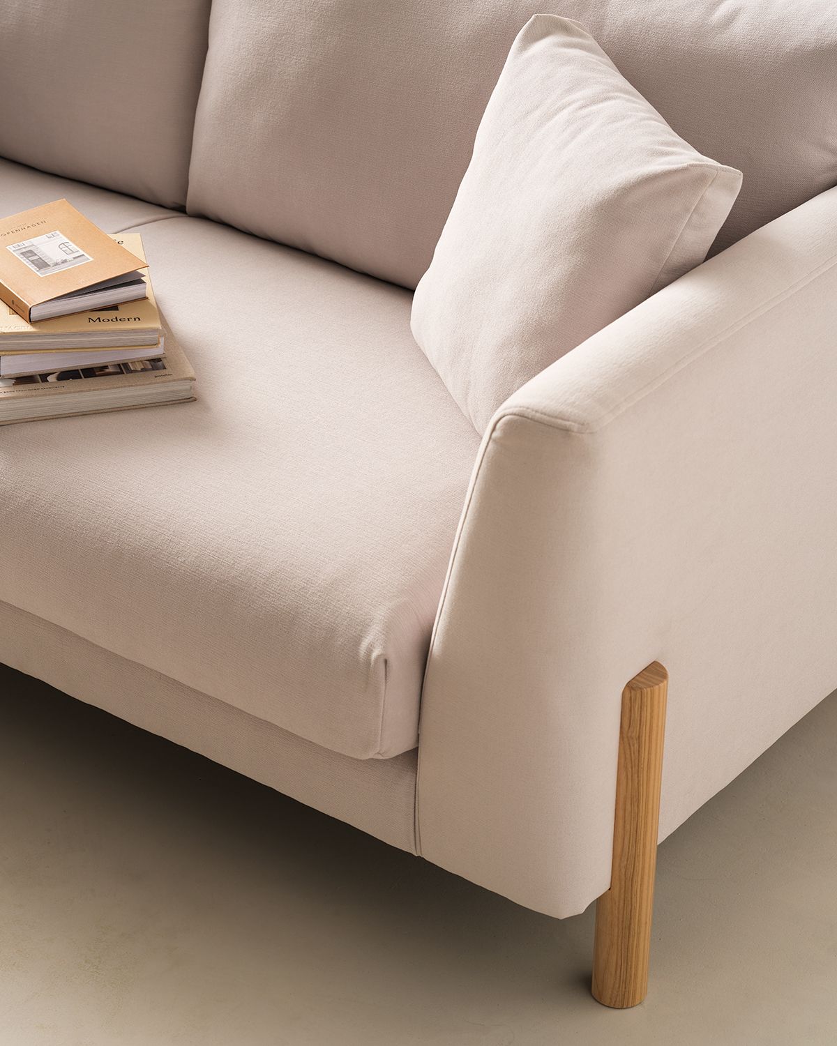 Close-up image of an off-white sofa seat and arm. There is a wooden leg in view. A matching off-white pillow and a stack of books are also on the seat.