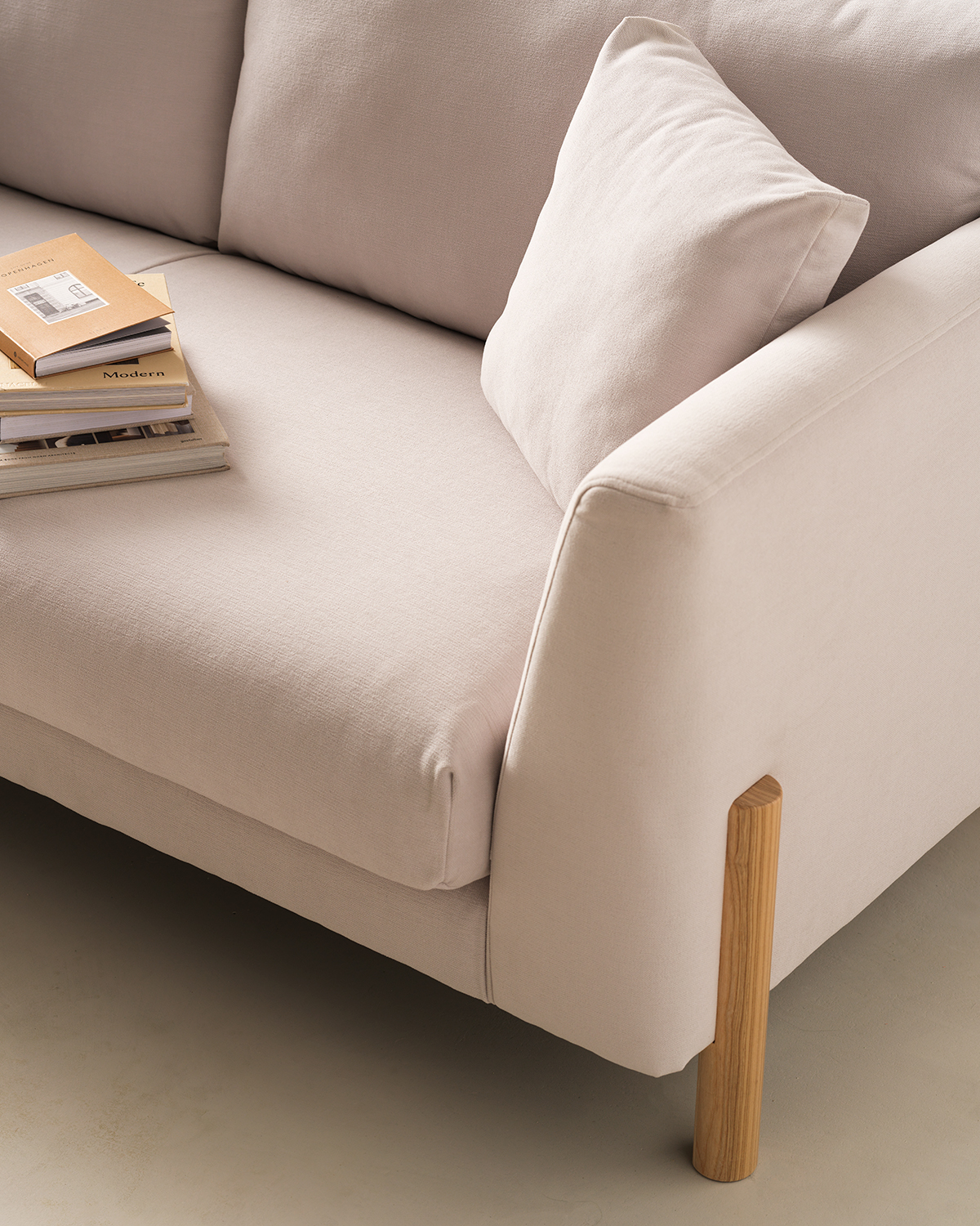 Close-up image of an off-white sofa seat and arm. There is a wooden leg in view. A matching off-white pillow and a stack of books are also on the seat.