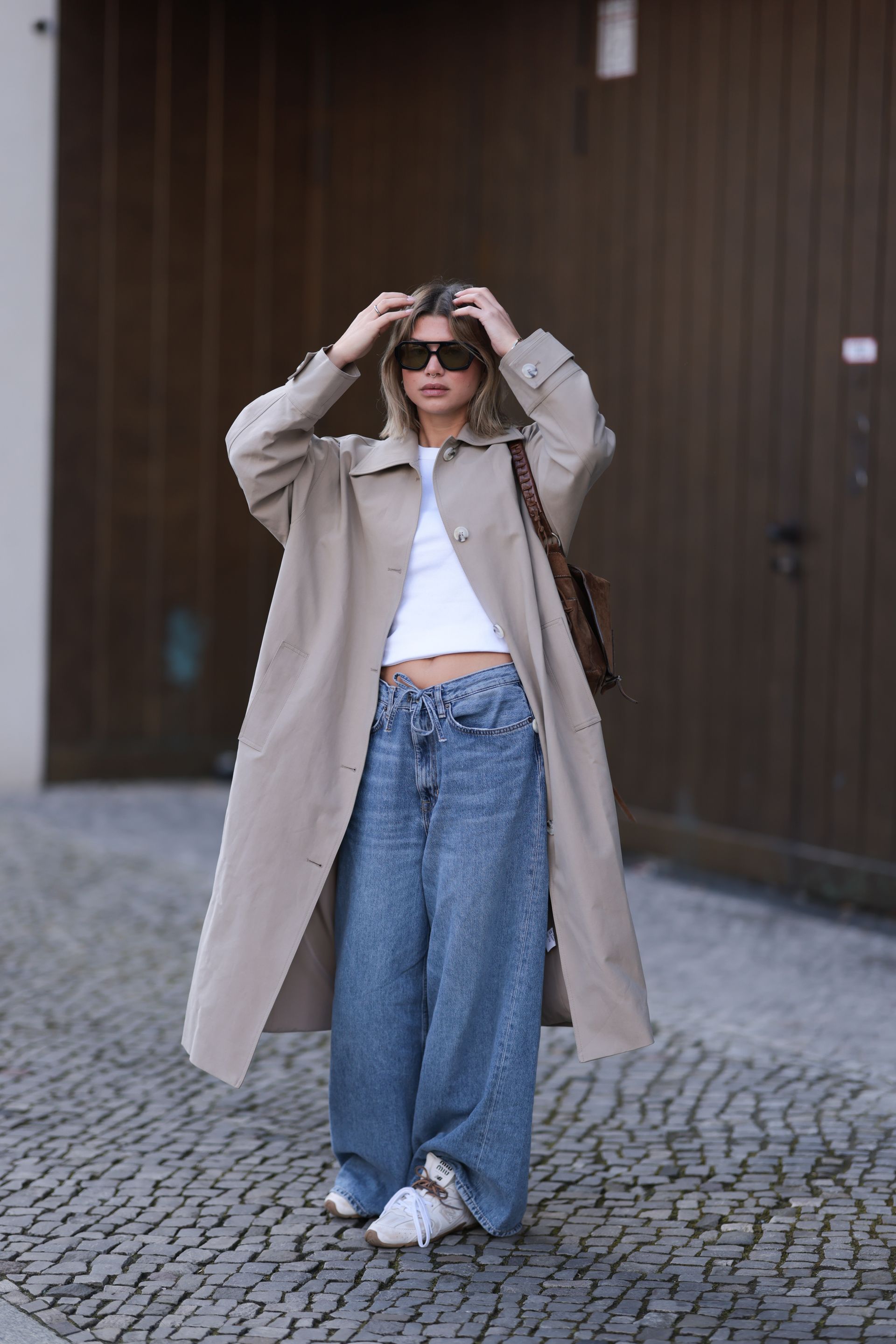 Fashion Person Sophia Geiss at Berlin fashion week. She wears the drawstring jeans trend and pairs them with a brown trench coat, white Miu Miu x New Balance trainers a white cropped tee and black sunglasses.