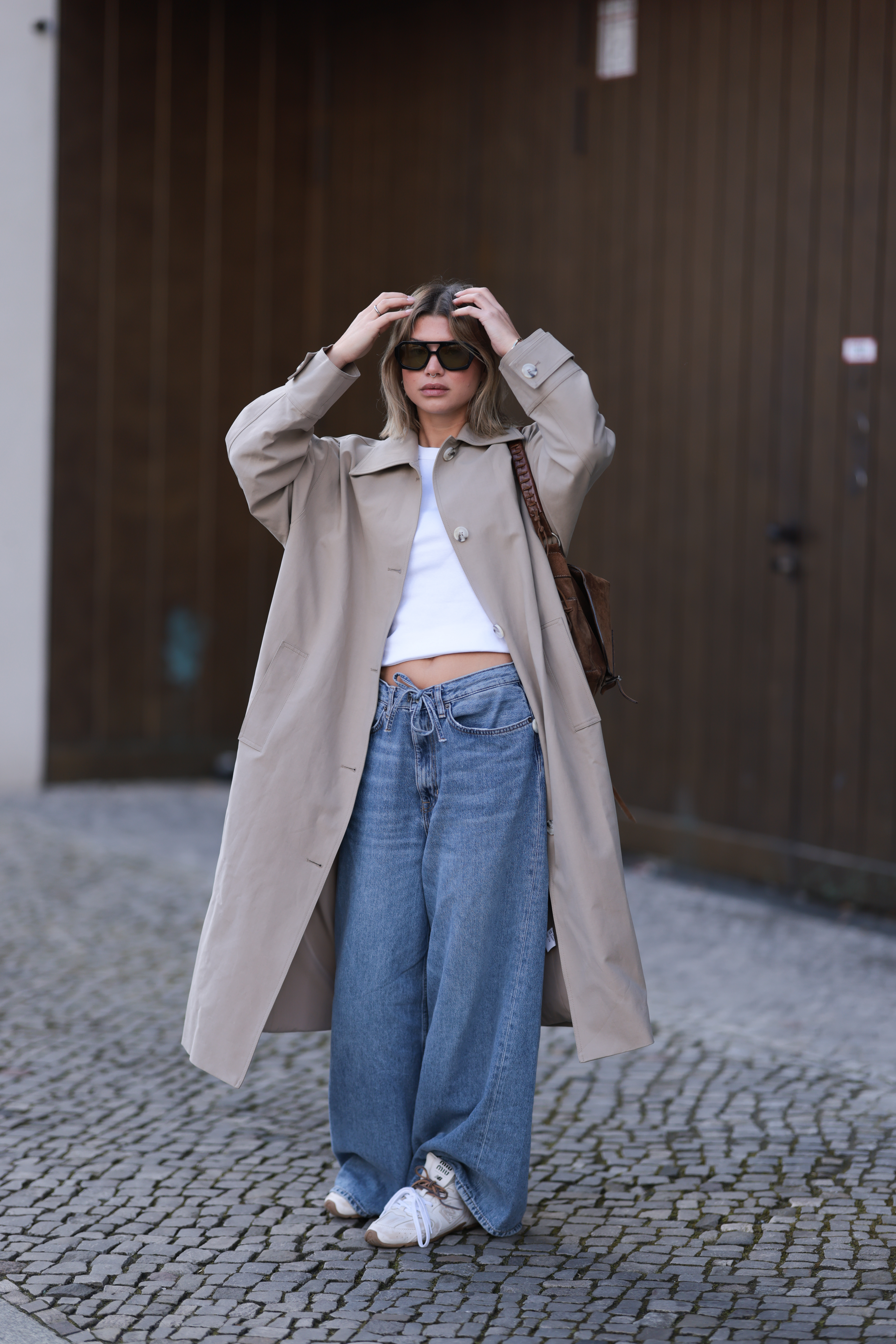 Fashion Person Sophia Geiss at Berlin fashion week. She wears the drawstring jeans trend and pairs them with a brown trench coat, white Miu Miu x New Balance trainers a white cropped tee and black sunglasses.