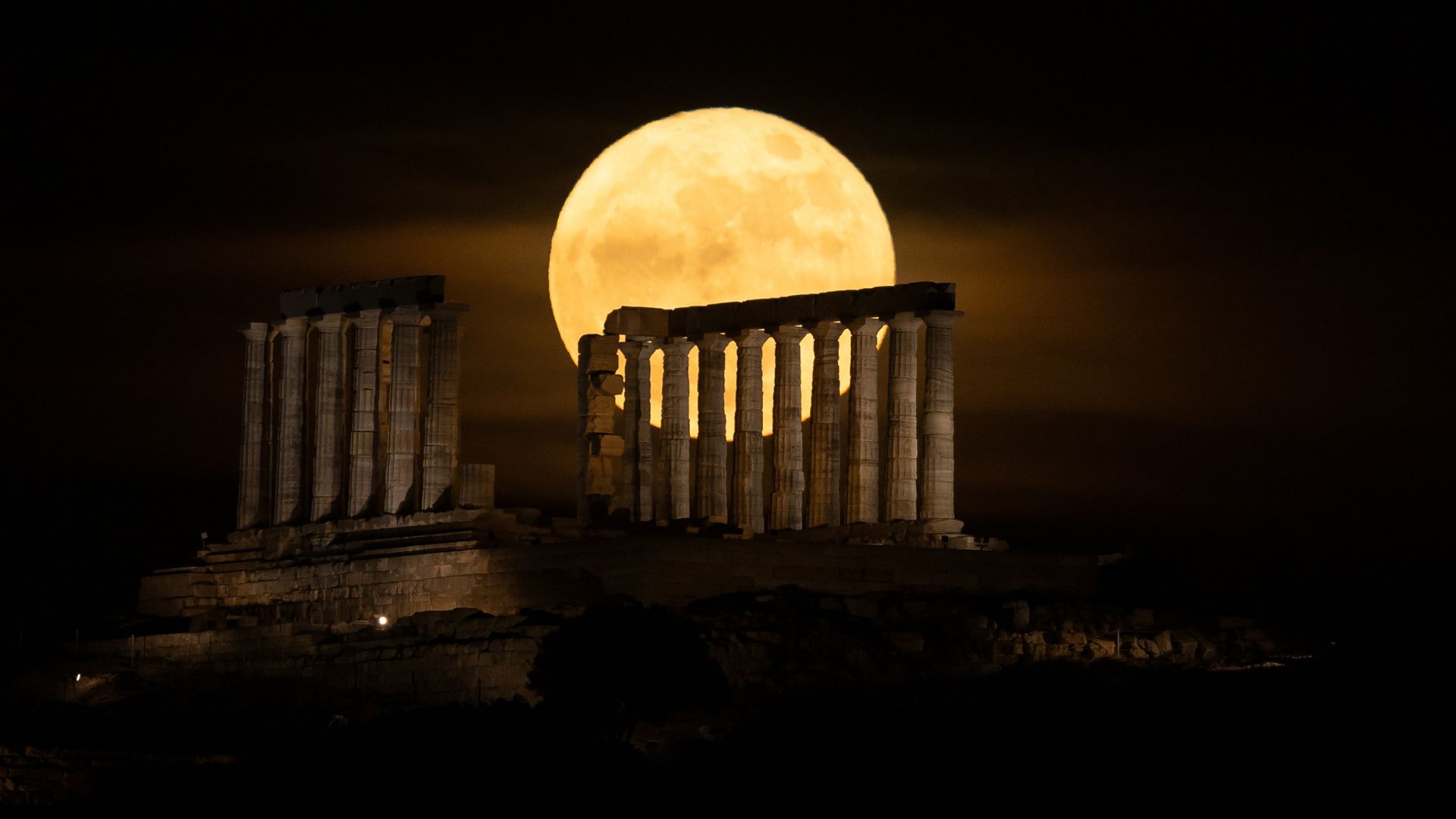 A yellow full moon glows in a dark sky behind the columns of an ancient ruined temple. 