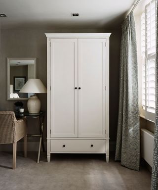 A white closet in a bedroom with large windows with white shutters and green and white patterned curtains.