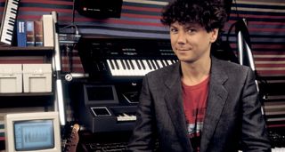 Jerry Harrison of Talking Heads in 1985, pictured with a grey blazer, red T-shirt, sitting in front of a bank of keyboards and a Telecaster