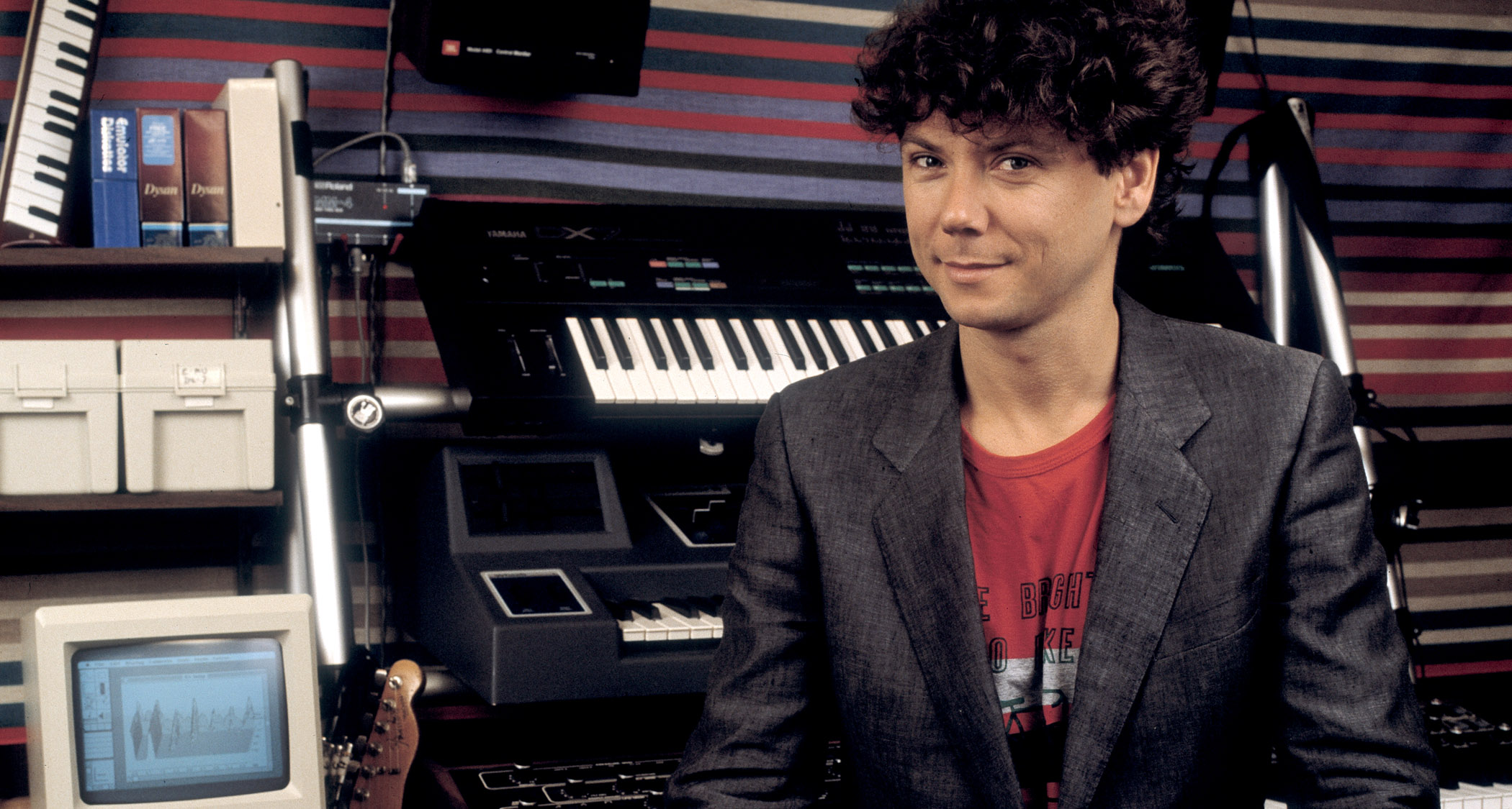 Jerry Harrison of Talking Heads in 1985, pictured with a grey blazer, red T-shirt, sitting in front of a bank of keyboards and a Telecaster 