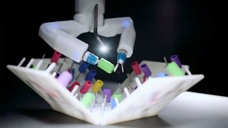 A screenshot of a video showing a surgical robot in action. A claw-like robot hand manipulates tiny O-rings, placing them onto tiny pegs.