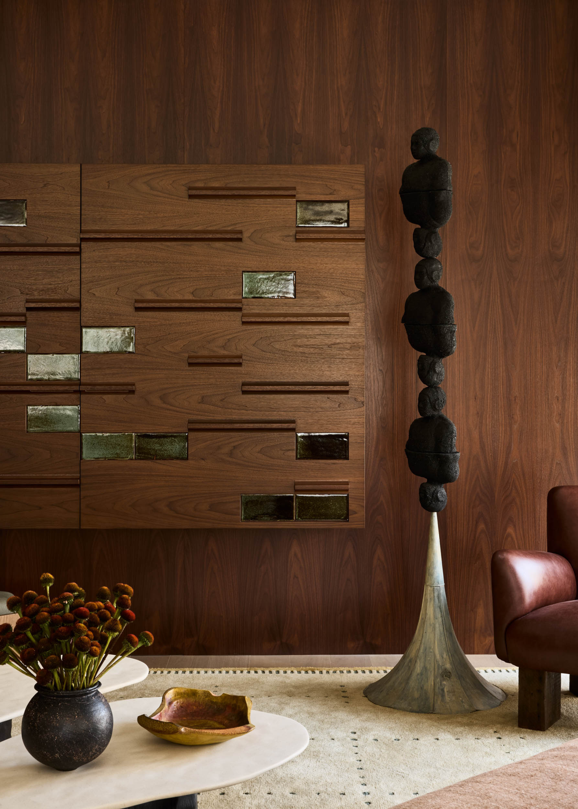 A living room with a structural black decor piece by a leather armchair and a wood cabinet with green glass inserts