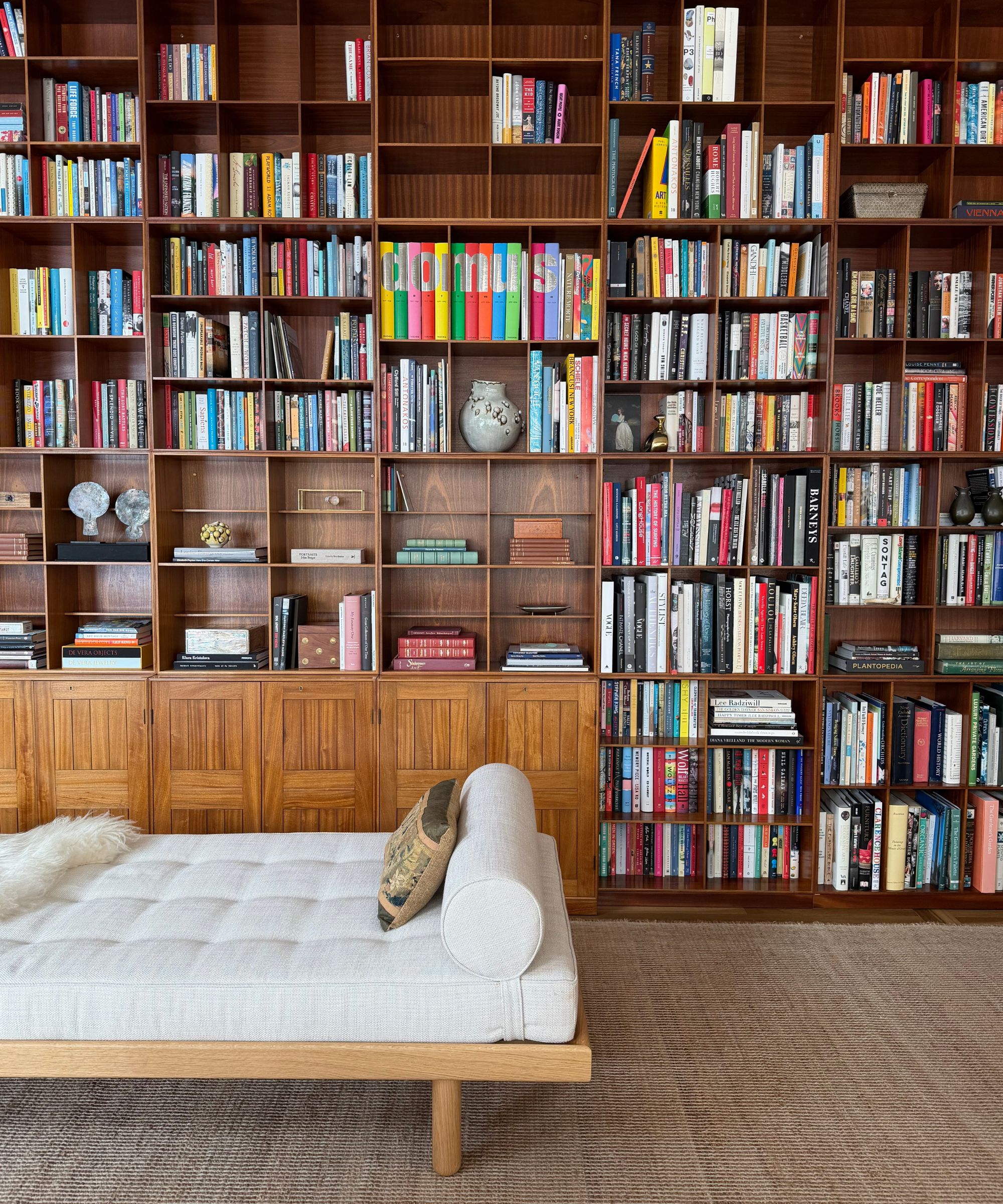 Wall-to-wall vintage teak bookcases filled with books and decorative objects, above paneled lower cabinets, styled behind a light upholstered daybed in a cozy library-style living room.