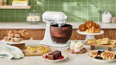 KitchenAid stand mixer with wooden bowl surrounded by cakes and bakes on a kitchen counter