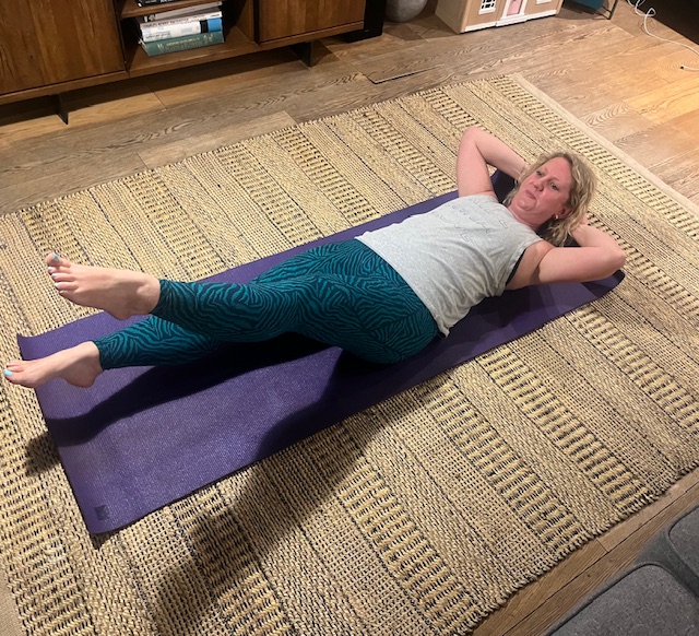 Kat Storr performs flutter kicks at home. She is reclining on a mat, arms behind her head and back pressed against the floor, with her left leg pointing upwards and her right leg hovering just above the floor.
