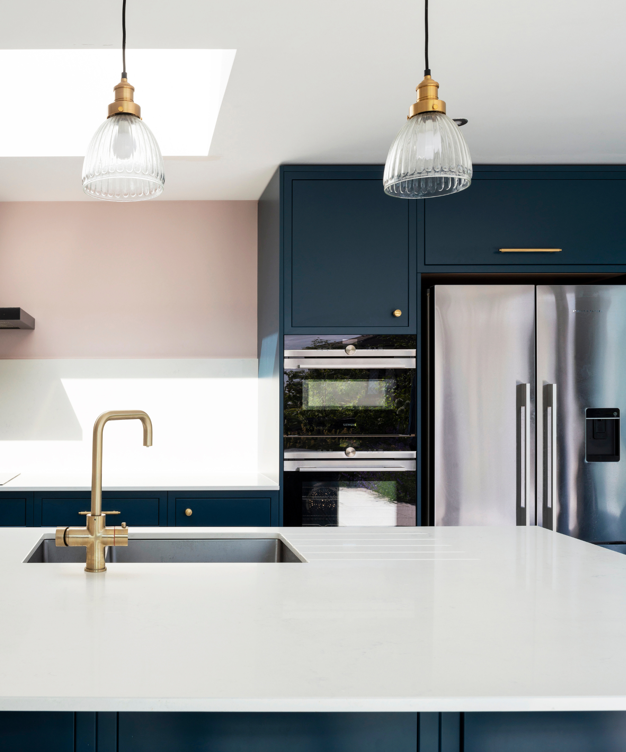 Close-up of the kitchen island which has a white worktop and a gold tap. Blue kitchen cabinets, silver fridge and pink half-wall on the other side with a roof light in the left hand corner