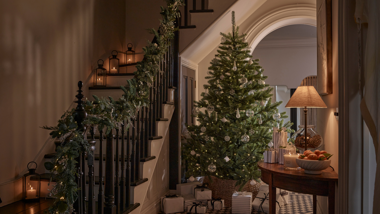 A staircase lit up by tealights with a winter garland running down the bannister. Next there is a christmas tree with silver decroations and silver presents at its feet.