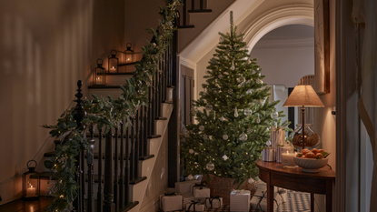 A staircase lit up by tealights with a winter garland running down the bannister. Next there is a christmas tree with silver decroations and silver presents at its feet.