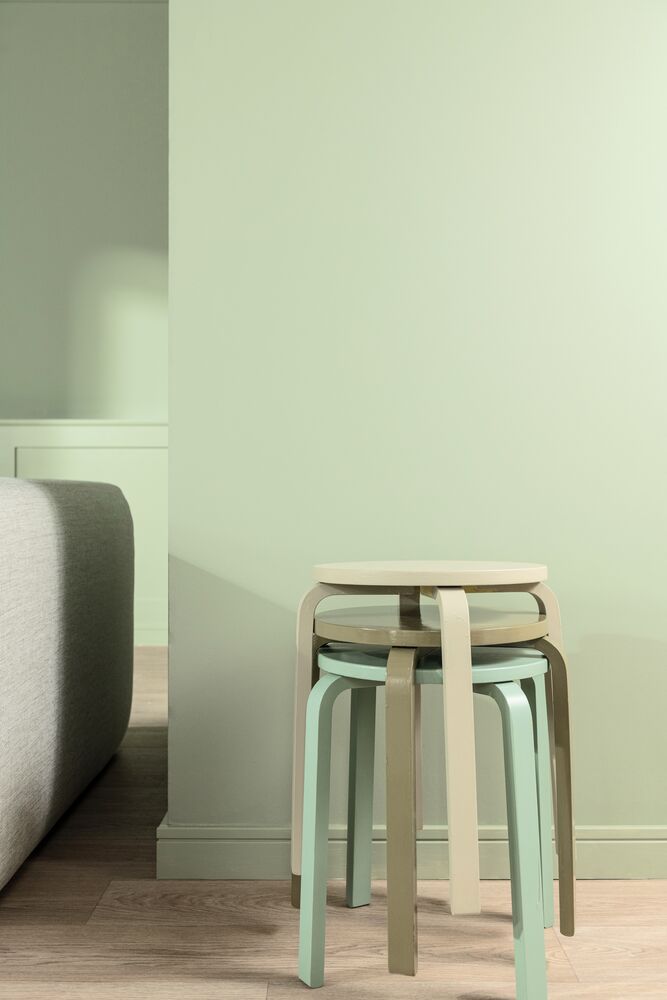 A sage green wall in a room with wooden floors. There are three green stools stacked on top of each other.