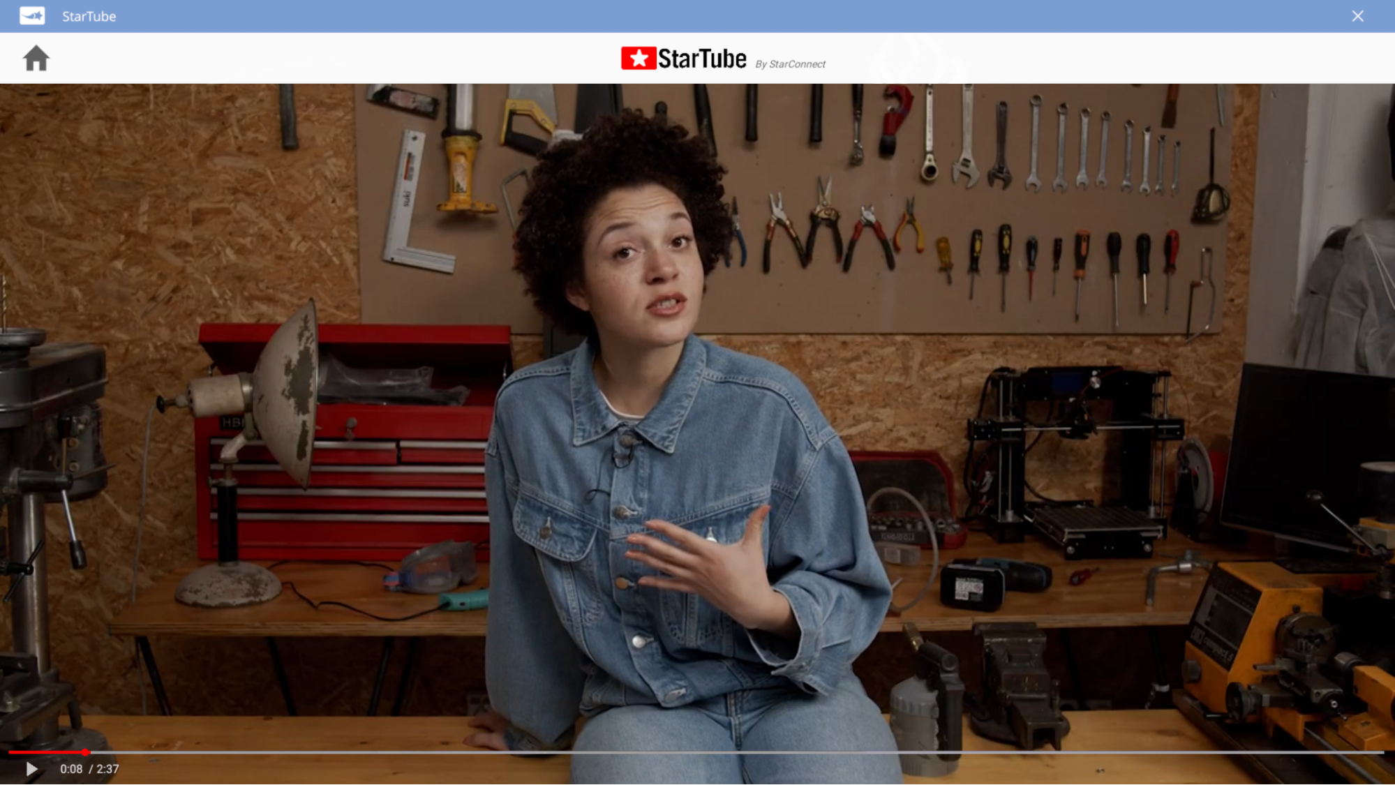 A screenshot of Next Space Rebel’s YouTube parody called StarTube. It’s a screencap of a woman wearing a denim jumpsuit talking as she sits on a workshop table. She is surrounded by tools.