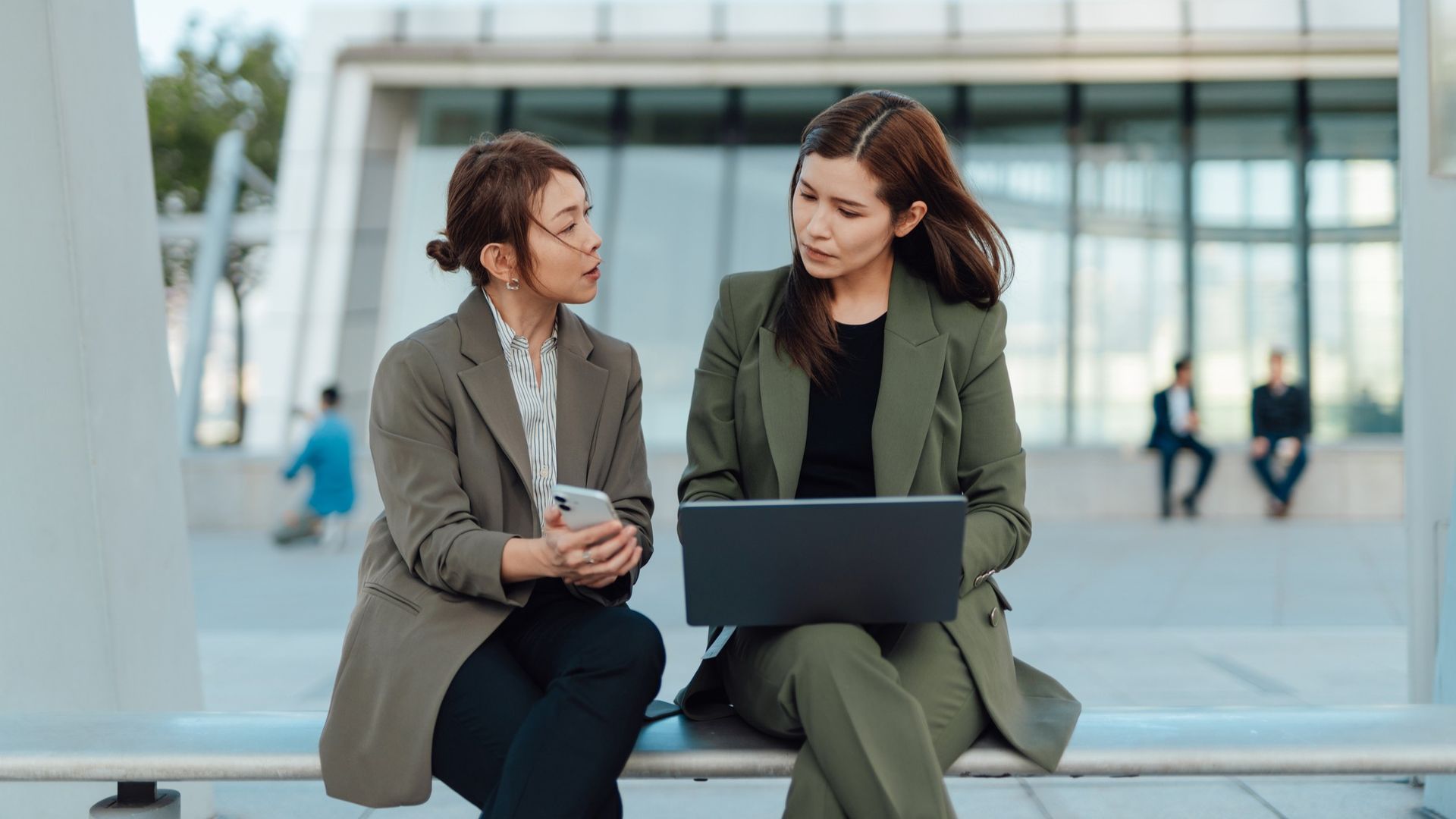 two women sitting outside an office building, one on a laptop looking at the other woman's smartphone screen