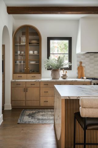 kitchen cabinetry with wood and arched cabinets. The arched cabinets have glass doors where plates and other essentials are placed. In the middle is a kitchen island with a marble countertop and bar stools.