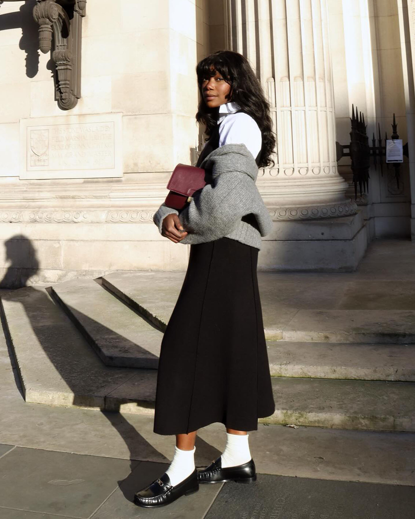 French fashion influencer Andi Mun poses on the streets of Paris in a white button-down shirt, gray sweater, burgundy bag, black skirt, white ankle socks, and black loafers