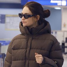 Lily James walking through Milan Malpensa Airport during Milan Fashion Week wearing a brown puffer coat and matching sweatpants, Prada sunglasses, and Prada's 2024 It bag, the buckle belt bag.