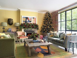 Colorful Christmas living room with yellow walls, green and blue couches, multi coloured rug, coffee table, garland above fireplace, decorated tree, artwork, lamps